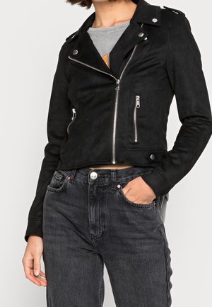 Person trägt eine schwarze Wildleder-Bikerjacke mit silbernen Reißverschlüssen und Knöpfen, kombiniert mit hoch taillierten schwarzen Jeans und einem grauen T-Shirt.