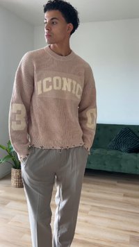 Vaaleanpunainen neulepaita, jossa on "ICONIC" vaalean beigen värillä, ribbikuviointi ja leikattu helma, yhdistettynä vaalean beigen värisiin housuihin.