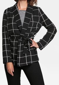 Svart kavaj med vit rutmönster, bälte i midjan, långa ärmar och sjalkrage, över en randig topp.