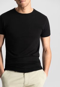Black short-sleeve t-shirt made of smooth fabric, featuring a round neckline. Worn with light-colored pants, hands in pockets.