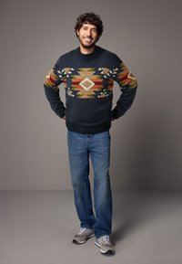 Gebreide trui in donker marineblauw met kleurrijke geometrische patronen. Gecombineerd met blauwe jeans en grijze hardloopschoenen. Heeft een geribde kraag.
