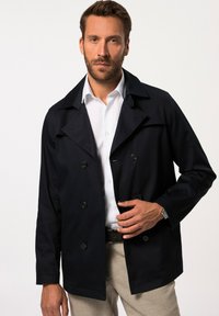 Abrigo azul marino de doble botonadura con cuello con muesca, cierre de botones y bolsillos laterales, combinado con una camisa blanca y pantalones beiges.