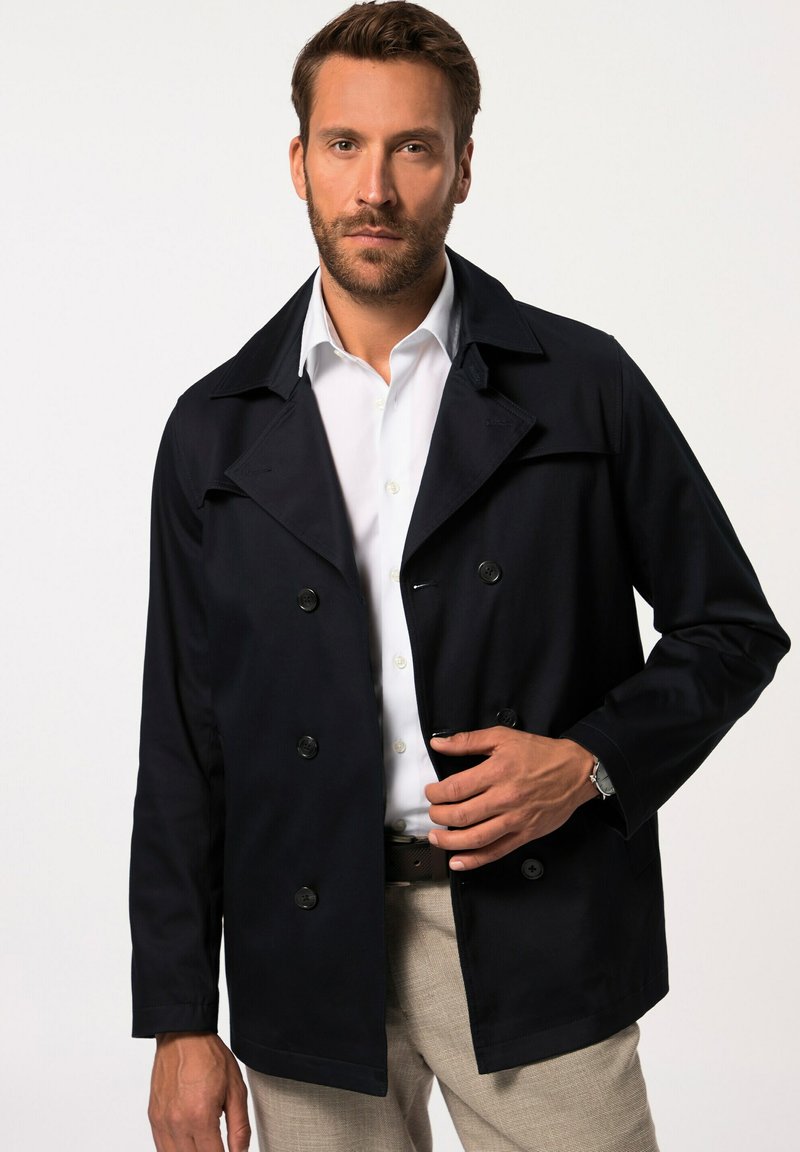 Abrigo azul marino de doble botonadura con cuello con muesca, cierre de botones y bolsillos laterales, combinado con una camisa blanca y pantalones beiges.