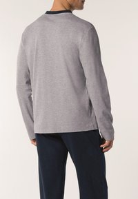 Uomo che indossa una camicia grigia a maniche lunghe con colletto nero e pantaloni blu scuro, visto da dietro su uno sfondo semplice.