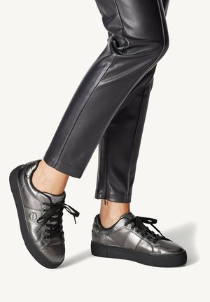 Metallisch graue Sneakers mit glatter Textur, runder Zehenpartie, schwarzen Schnürsenkeln und kontrastierender schwarzer Sohle; kombiniert mit eng anliegenden schwarzen Lederhosen.