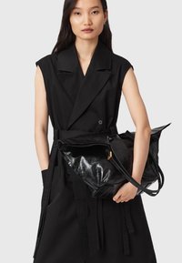 Donna che indossa un trench senza maniche di colore nero mentre tiene una grande borsa tote in pelle nera lucida contro uno sfondo grigio.