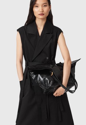 Donna che indossa un trench senza maniche di colore nero mentre tiene una grande borsa tote in pelle nera lucida contro uno sfondo grigio.