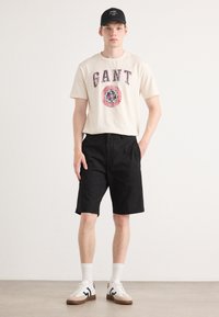 Beige cotton t-shirt with graphic print, black shorts, white socks, and cream sneakers with dark accents. Man standing with one hand in pocket.