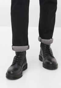 Stivali neri in pelle con lacci, parte superiore texturizzata e suola in gomma spessa. I pantaloni presentano un orlo piegato con un motivo a pois bianco e nero.