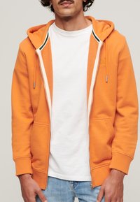 Orange zip-up hoodie med en snorhætte, frontlommer og ribbede manchetter, båret over en hvid t-shirt og lyseblå jeans.