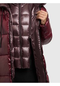 Bordinės spalvos pūsta striukė su blizgiu, siuvinėtu paviršiumi ir gobtuvu. Užsegama u�žtrauktuku, matoma vidinė pamušalas ir elastinės rankogaliai, užtikrinantys artimą prigludimą.