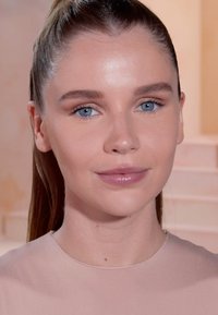 Portret van een persoon met recht, lang haar en een lichtgekleurde top. Kenmerken zijn een lichte huid, blauwe ogen en neutrale make-up.