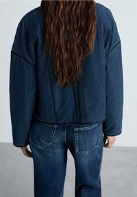 Veste courte en denim foncé avec des coutures verticales et des manches larges, associée à un jean taille haute avec poches arrière. Tissu texturé.