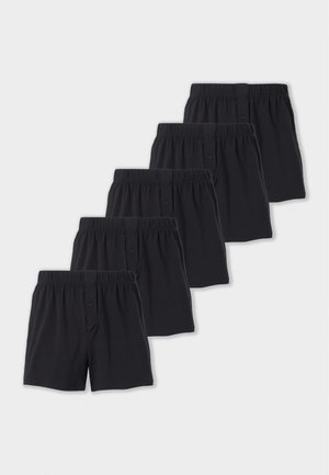 Homme torse nu, debout pieds nus, les bras croisés, portant un boxer noir sur un fond clair uni.