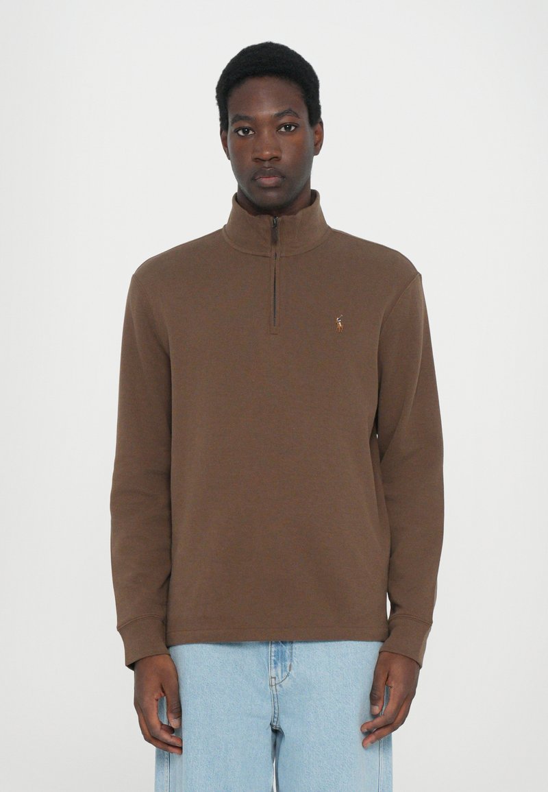 Bruine zip-up fleece pullover met een hoge kraag, voorzien van lange mouwen en een klein geborduurd logo op de borst. Gekleed met lichtblauwe jeans.