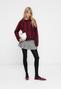 Bordeauxkleurige gebreide trui met zigzagpatroon, grijze geplooide rok, zwarte panty en rode balletschoenen. Witte tas met zwarte accenten.