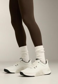 Białe buty sportowe z teksturowanej tkaniny, szare akcenty i czarne podeszwy, noszone z białymi prążkowanymi skarpetkami i brązowymi legginsami.
