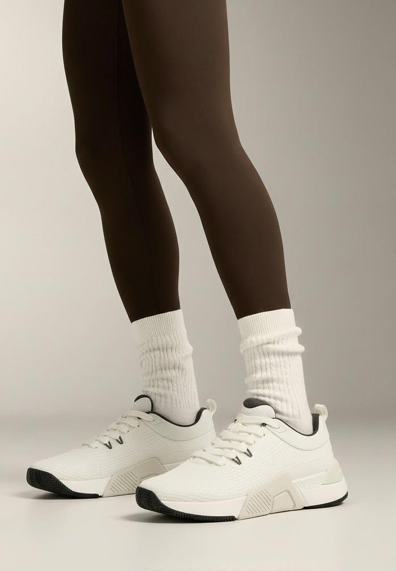 Białe buty sportowe z teksturowanej tkaniny, szare akcenty i czarne podeszwy, noszone z białymi prążkowanymi skarpetkami i brązowymi legginsami.