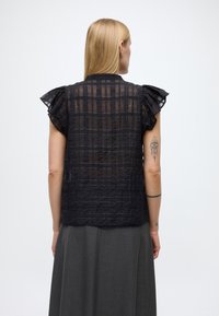 Svart, gjennomsiktig topp med teksturert rutemønster og volangerm. Har standardkrage og en avslappet passform, kombinert med et grått skjørt.