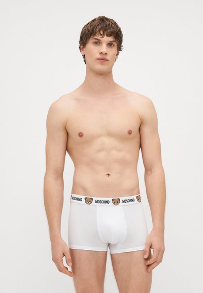 Rapaz jovem com cabelo castanho curto, vestindo boxers brancos com o logótipo Moschino e estampa de urso no cós, de pé contra um fundo simples.