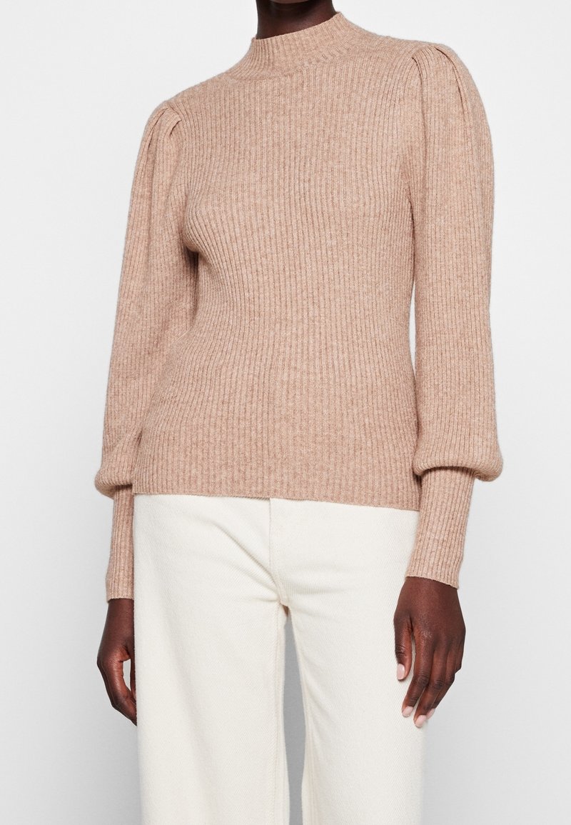 Femme portant un pull en maille côtelée beige avec des manches longues bouffantes et un pantalon blanc sur un fond uni.