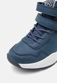 Zapatilla de cuero azul con superficie texturizada, suela de goma blanca, tres correas de cierre y cordones en contraste con patrones rojos y blancos.