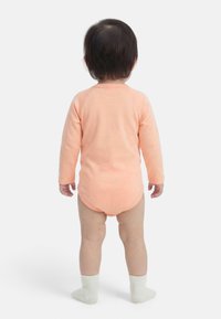 Νήπιο που στέκεται με την πλάτη προς τα έξω, φοράει ένα ανοιχτό ροδακινί φορμάκι με μακριά μανίκια και λευκές κάλτσες, μπροστά σε απλό λευκό φόντο.