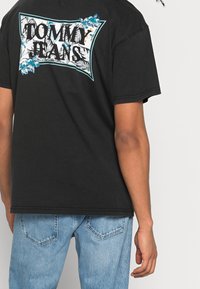 Camiseta de algodón negra con un diseño floral colorido y el texto “TOMMY JEANS” en la parte posterior, combinada con jeans de denim azul claro.