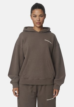 Donna con capelli scuri raccolti che indossa una felpa marrone oversize e pantaloni abbinati con piccoli loghi bianchi sul petto e sulla coscia.
