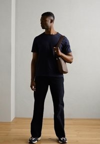 Schwarzes Baumwoll-T-Shirt, dunkle Jeans und sportive Schuhe. Braune Umhängetasche. Stehend gegen eine hellgraue Wand auf einem Holzboden.