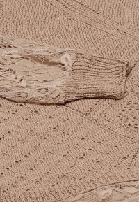 Beige kötött pulóver textúrált mintákkal, csipkés részletekkel a ujjaknál és bordás mandzsettákkal, amely egy puha, nyitott szövésű designt mutat be.
