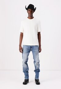 Weiße gestrickte Kurzarmbluse mit vertikaler Rippung, hellblaue verwaschene Jeans und schwarzen Cowboyhut, ergänzt durch schwarze Lederstiefeletten.