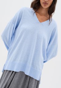 Frau in lockerer, hellblauer V-Ausschnitt-Pullover und grauem, plissiertem Rock vor einem schlichten weißen Hintergrund.