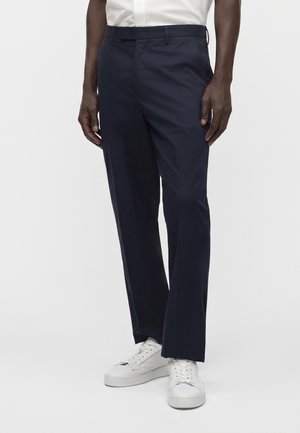 Homme portant un pantalon droit bleu marine foncé et des baskets blanches, debout devant un fond blanc uni.