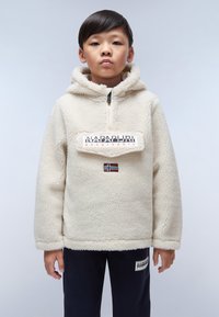 Ung person som bär en krämfärgad fleecehoodie med Napapijri-logotyp och norsk flagga, samt mörka byxor med en matchande logotypapplikation.