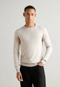 Beige hosszú ujjú pulóver kerek nyakkivágással, bordás mandzsettával és szegéllyel, a mellkason egy kis sárga logóval, sötét nadrággal viselve.