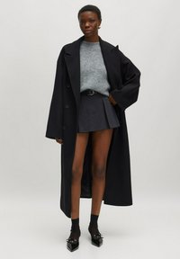 Zwarte lange jas over een lichtgrijs sweater, gecombineerd met een donkere geplooide rok. Draagt zwarte enkelsokken en puntige schoenen. Glad textuur.