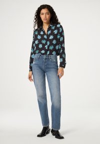 Schwarze Bluse mit floralem Muster in Blau, kombiniert mit hellblauen Jeans. Das Outfit wird mit schwarzen Knöchelboots abgerundet.