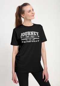 Mladá žena s dlouhým culíkem je oblečena v černém tričku s nápisem "Journey 1983 Faithfully" a s křídly emblémem, dívá se nahoru a doprava.