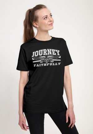 Mladá žena s dlouhým culíkem je oblečena v černém tričku s nápisem "Journey 1983 Faithfully" a s křídly emblémem, dívá se nahoru a doprava.