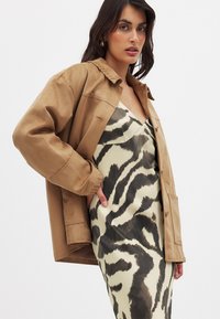 Veste en daim marron avec un col, portée sur une robe crème avec un motif zèbre noir, mettant en valeur un design et des textures élégants.