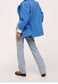 Chaqueta oversized de mezcla de lana azul con un amplio cuello, puños de punto crema y bolsillos laterales, combinada con jeans rectos de mezclilla azul claro y zapatos marrones.