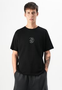 Junger Mann in schwarzem T-Shirt mit weißem Design eines Halbmonds und einer Wolke, steht mit den Händen in den Taschen der dunklen Hose vor einem weißen Hintergrund.