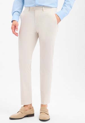 Homme portant un pantalon slim beige clair, une chemise bleu clair et des mocassins en daim beige, avec une main dans la poche sur fond blanc.