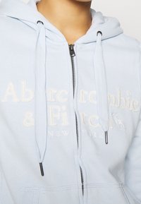 Jasnoniebieska bluza z kapturem na zamek z trokami, z wyższym białym napisem "Abercrombie & Fitch New York" oraz małym logiem jelenia na klatce piersiowej.