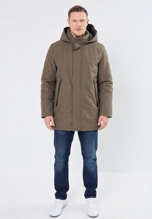Hombre con abrigo de invierno verde oliva con capucha, vaqueros azules y zapatillas blancas, de pie sobre un fondo liso y claro.