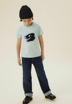 Vaaleansininen t-paita, jossa on musta ja tummansininen graafinen kuvio, yhdistetty tummiin farkkuihin ja mustiin kenkiin. Lapsella on päässään musta neulottu pipo.