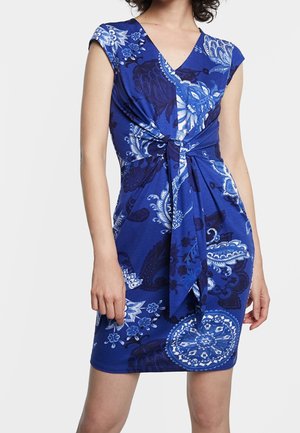 Niebieska sukienka do kolan z białymi i granatowymi kwiatowymi wzorami, dekoltem w serek, krótkimi rękawkami i wiązaniem z przodu w talii, noszona przez osobę.