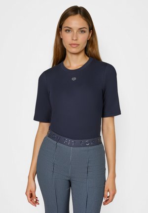 Young woman with long brown hair wearing a fitted navy short-sleeve top and blue patterned high-waist pants with a branded waistband.