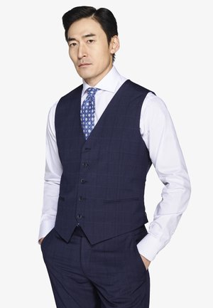 Mężczyzna w granatowej kratkowanej kamizelce i spodniach, białej koszuli wizytowej oraz niebieskim krawacie w wzory, stojący z rękami w kieszeniach.